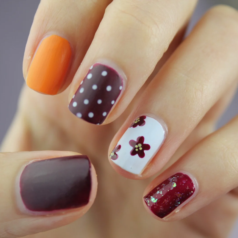 Mano con cinco uñas pintadas en naranja, ciruela, lunares blancos, diseño floral y purpurina roja.