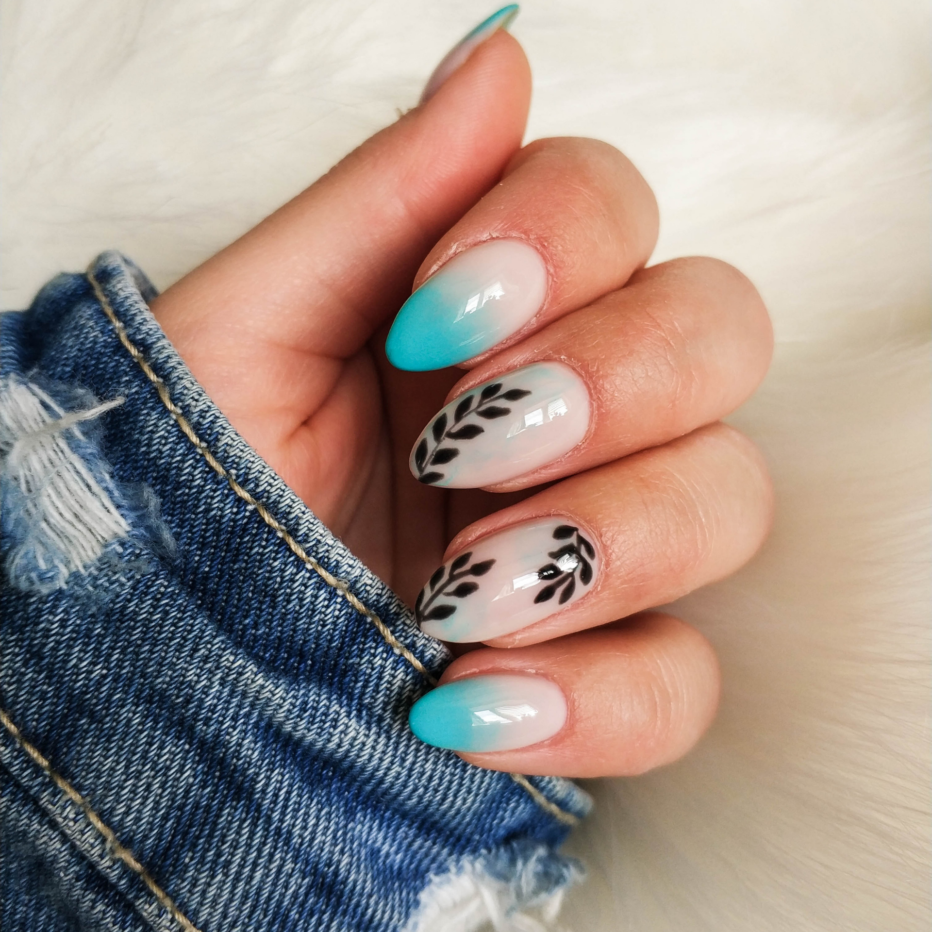 Mano con uñas ovaladas, algunas con diseño de hojas color verde azulado y otras con hojas negras.