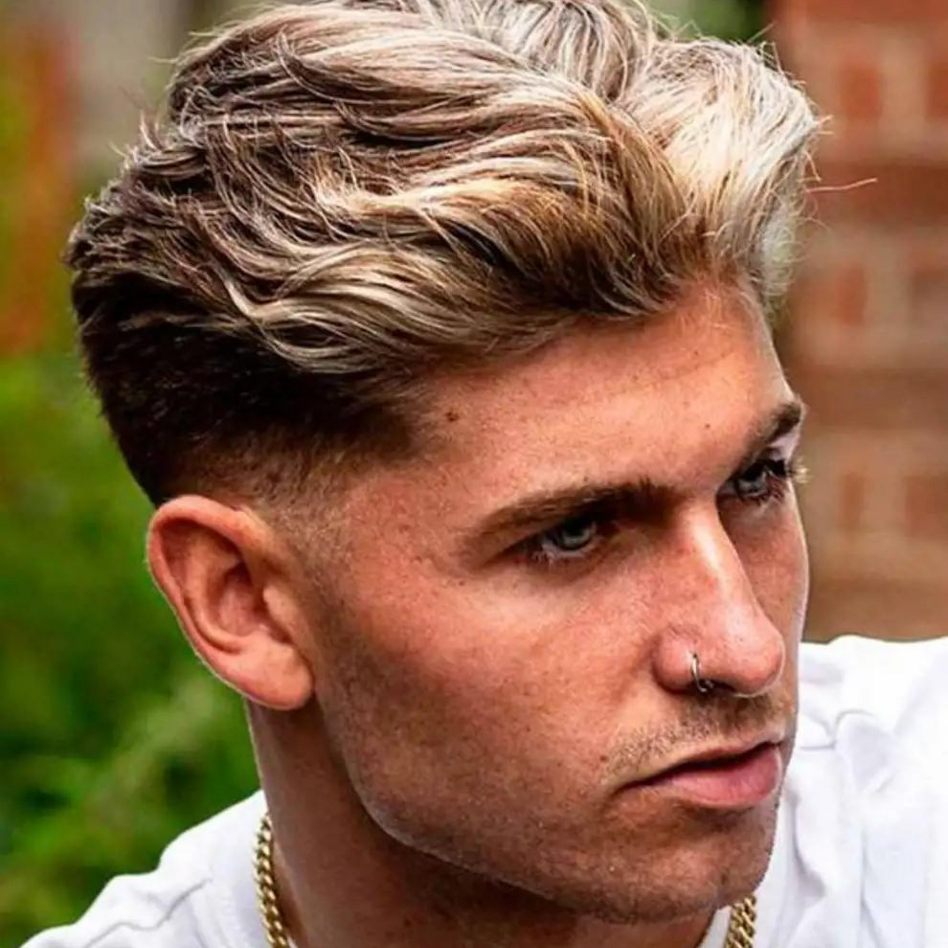 Primer plano de un hombre con el pelo con mechas y un piercing en la nariz mirando hacia un lado al aire libre.