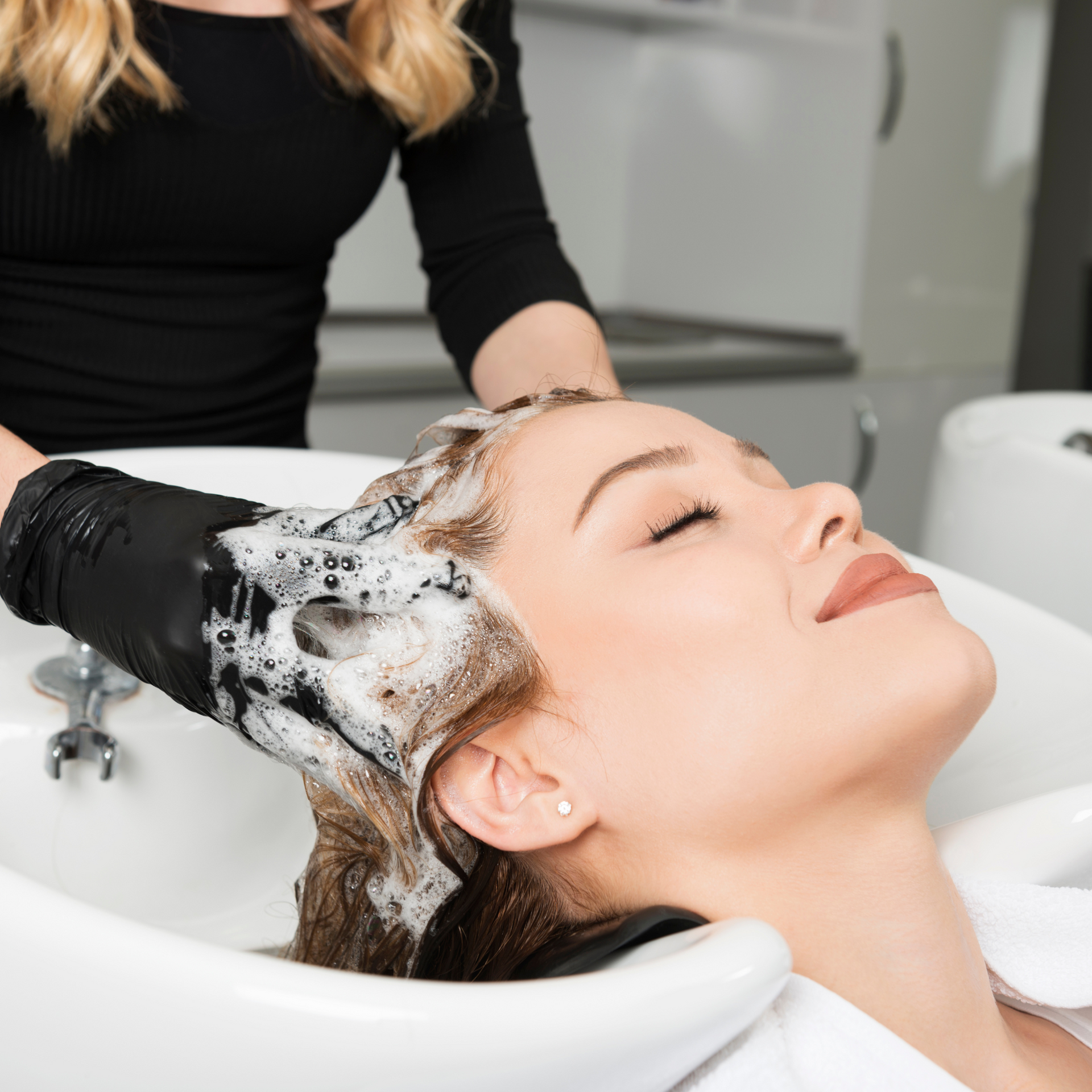 Una peluquera lava el cabello de una clienta en el lavabo de la peluquería, con espuma de champú, mientras la clienta está cómodamente recostada.