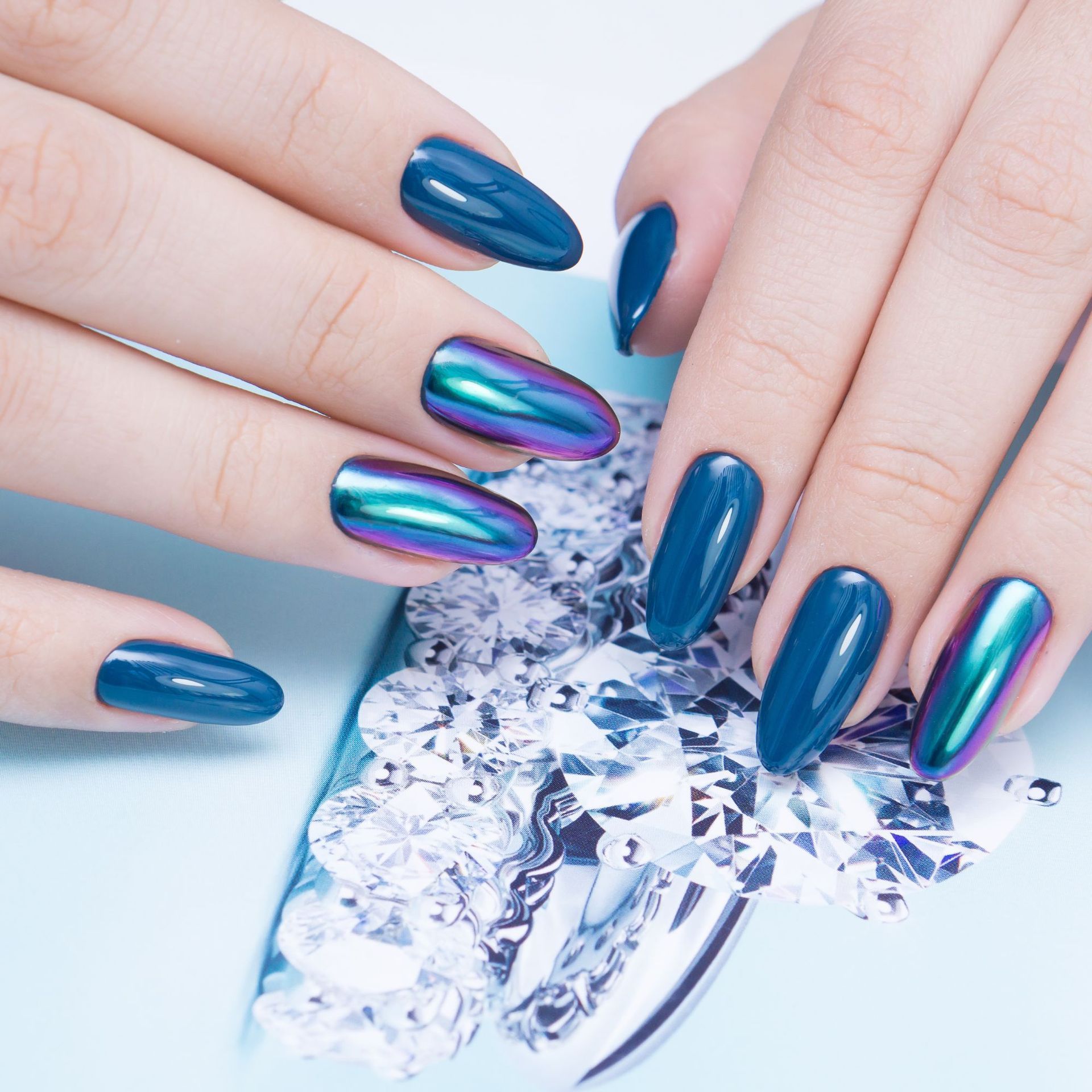 Manos con uñas largas de color verde azulado y púrpura iridiscente sosteniendo una gema facetada de plata.