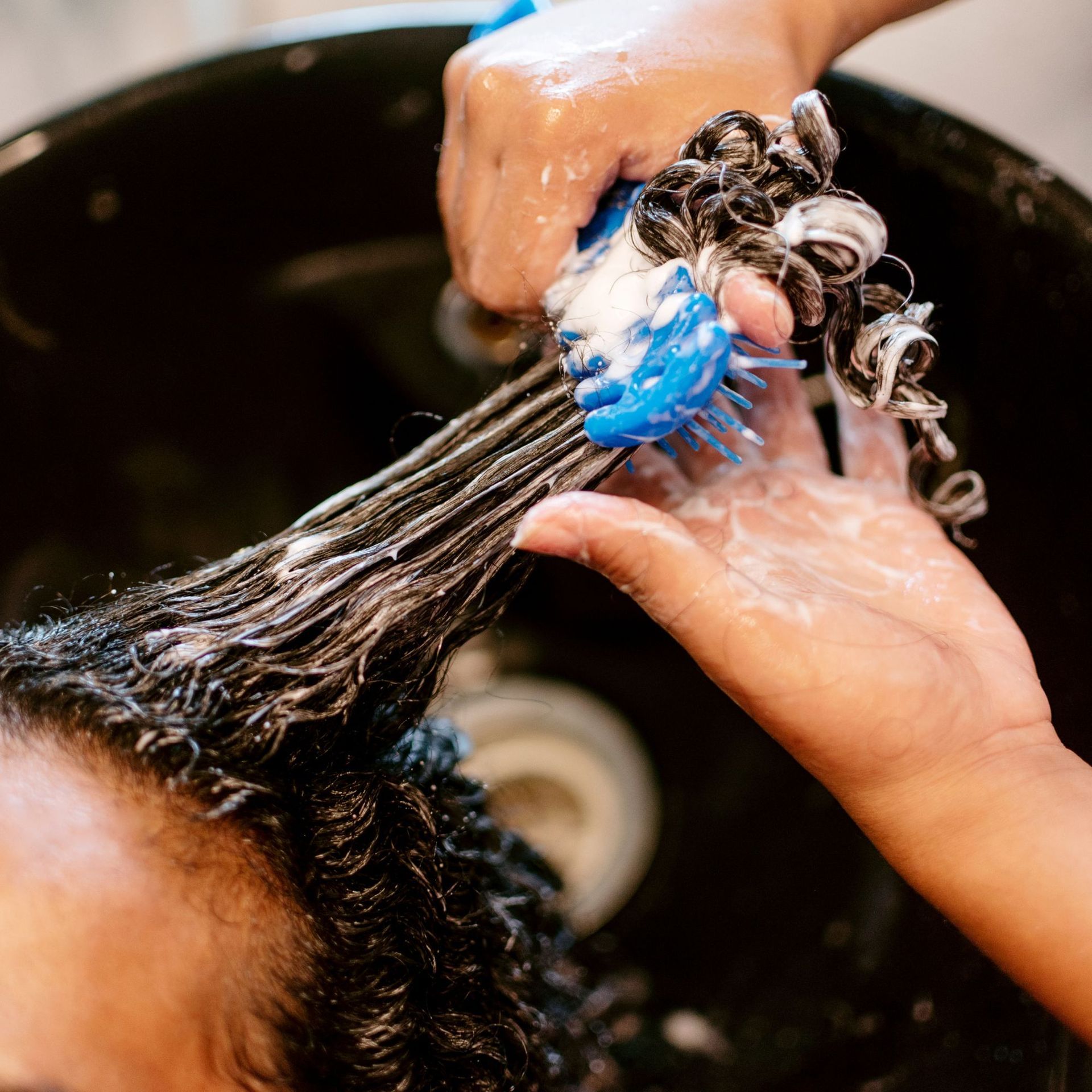 El cabello se está lavando en un lavabo, mientras unas manos masajean el champú a través del cabello oscuro trenzado.