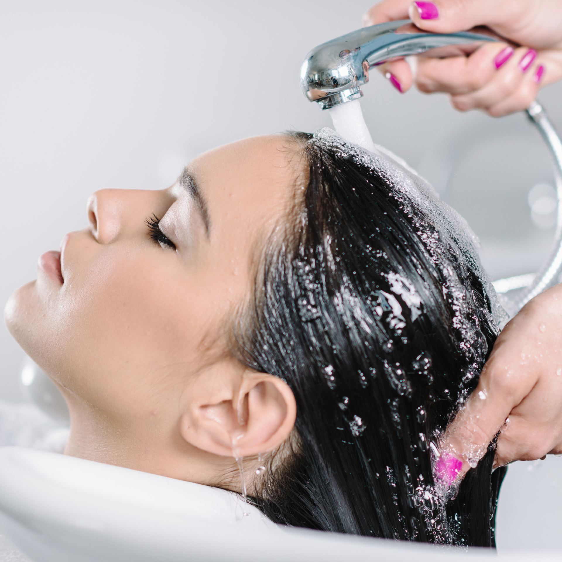 Mujer recostada en el lavabo de un salón de belleza mientras el agua enjuaga su cabello oscuro.