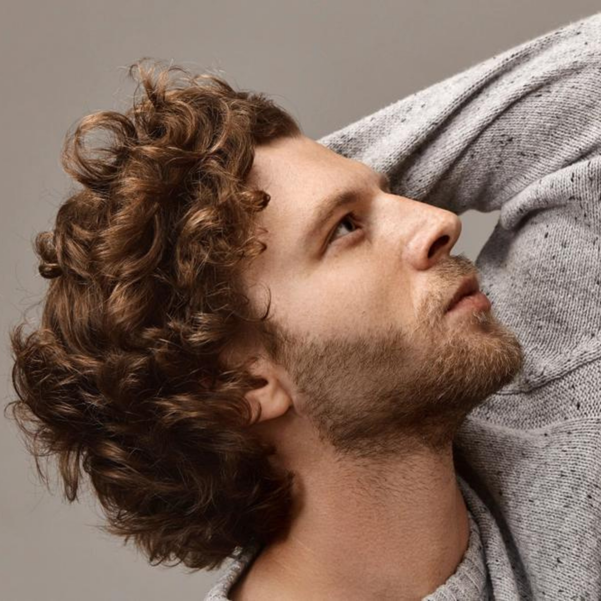 Hombre de pelo rizado de perfil, apoyado en un cojín gris, mirando hacia arriba con expresión pensativa.