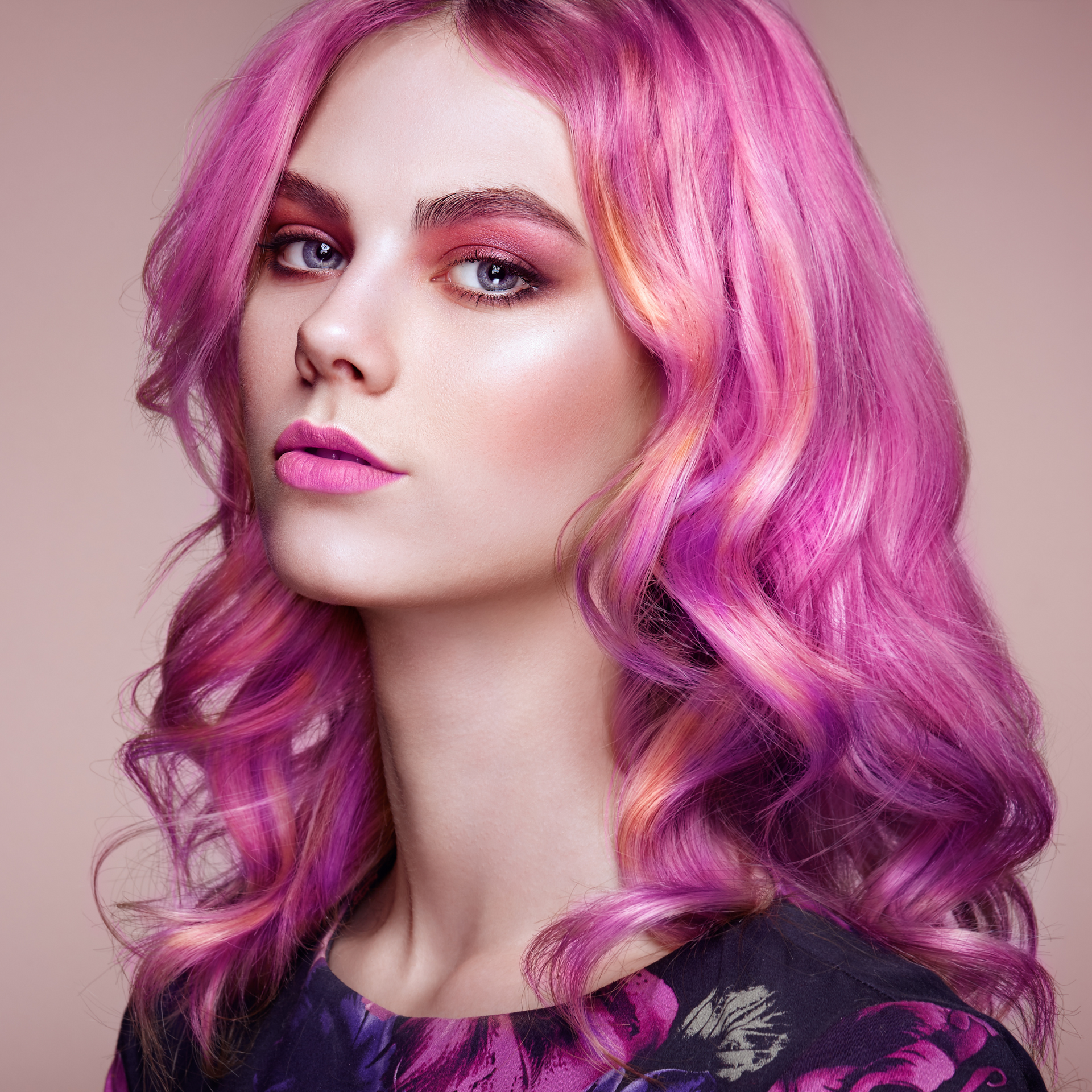 Retrato de una persona con cabello ondulado rosa brillante y maquillaje, mirando por encima del hombro contra un fondo liso.