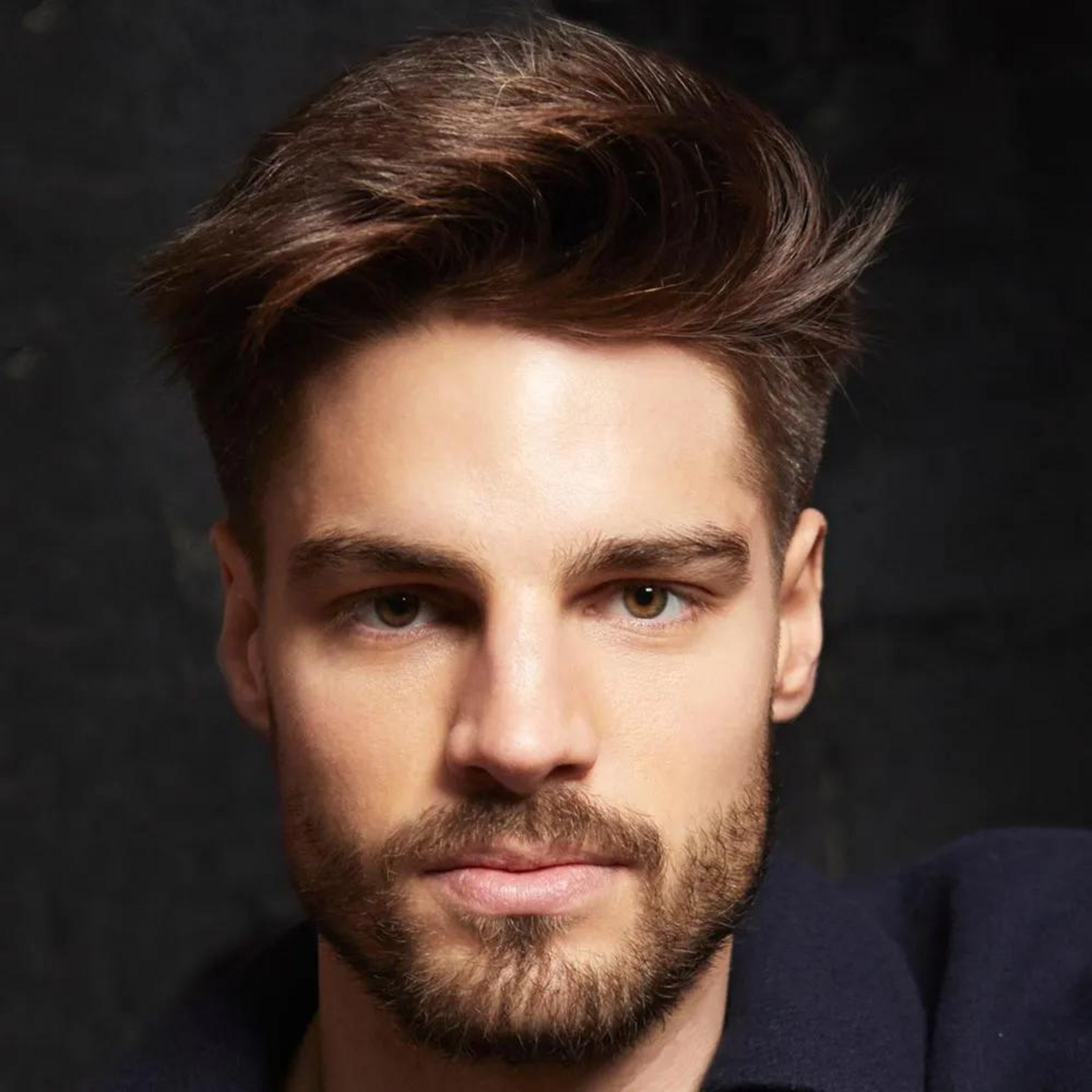 Retrato de estudio de un hombre con cabello castaño peinado y barba sobre un fondo oscuro.