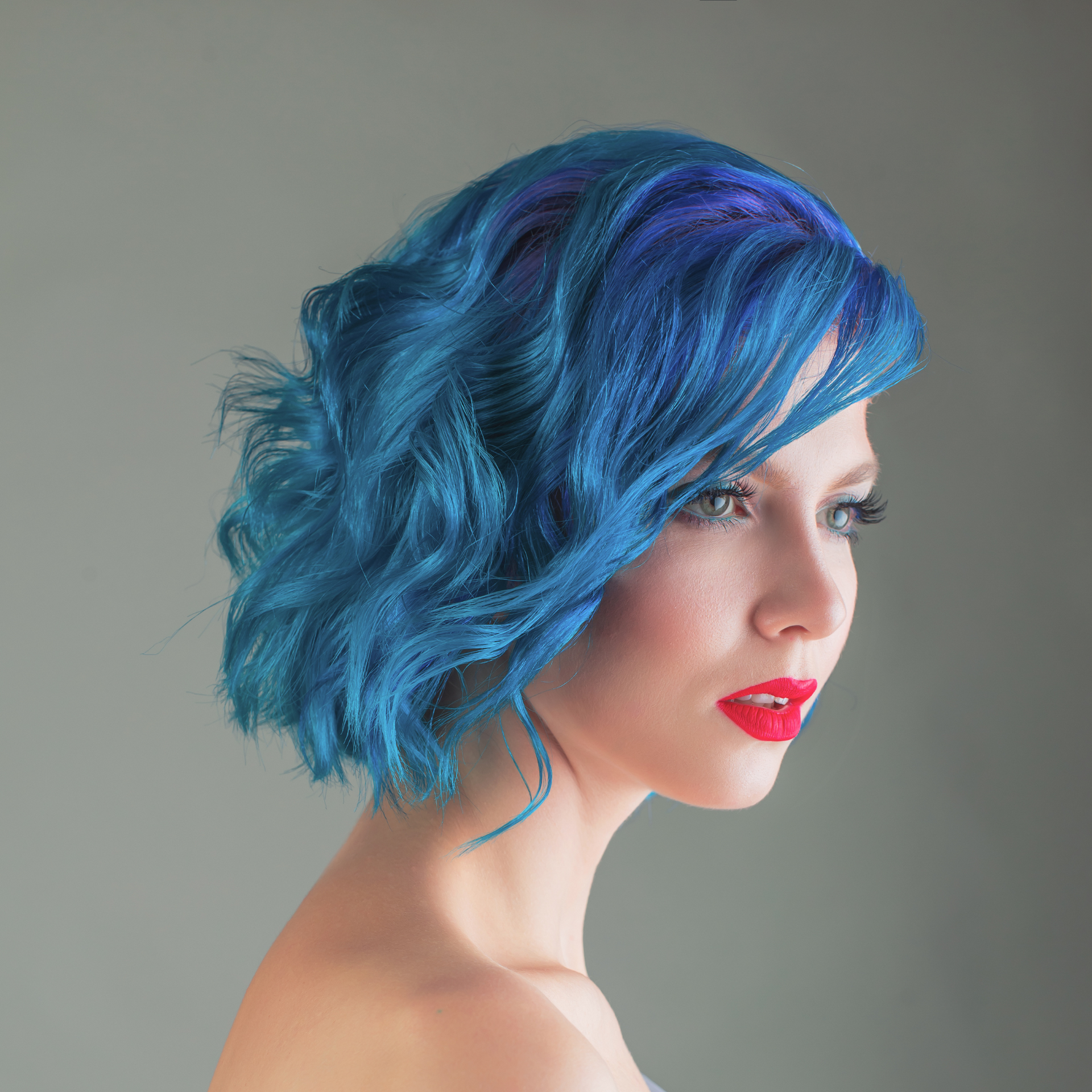 Perfil de una persona con cabello corto azul brillante y lápiz labial rojo sobre un fondo gris.