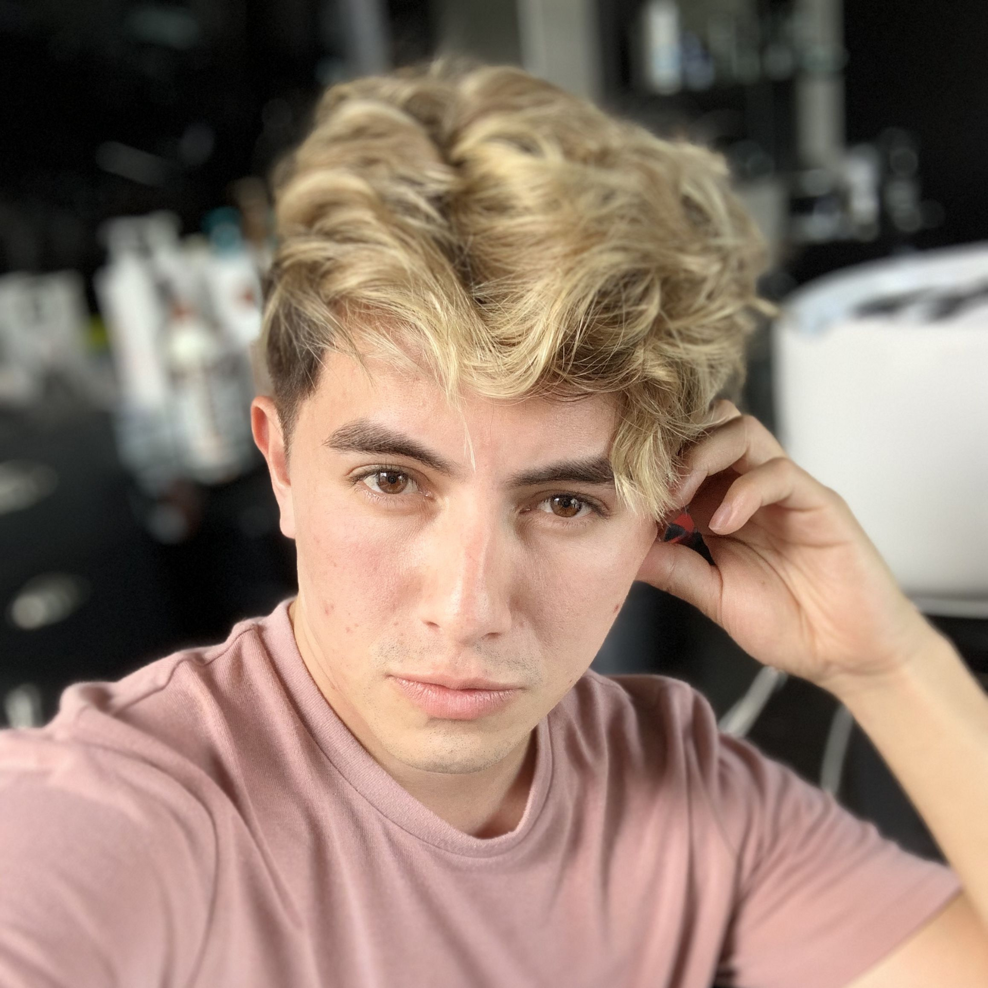 Persona joven con cabello rubio ondulado y camisa rosa, posando en interiores con una mano junto a la oreja.
