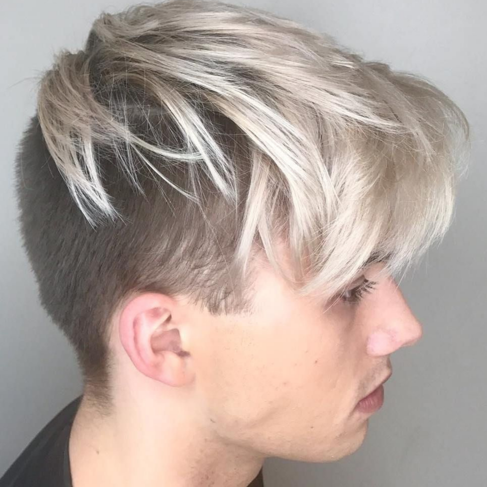 Vista de perfil de una persona con cabello rubio platino corto y rapado lateral oscuro sobre un fondo gris liso.