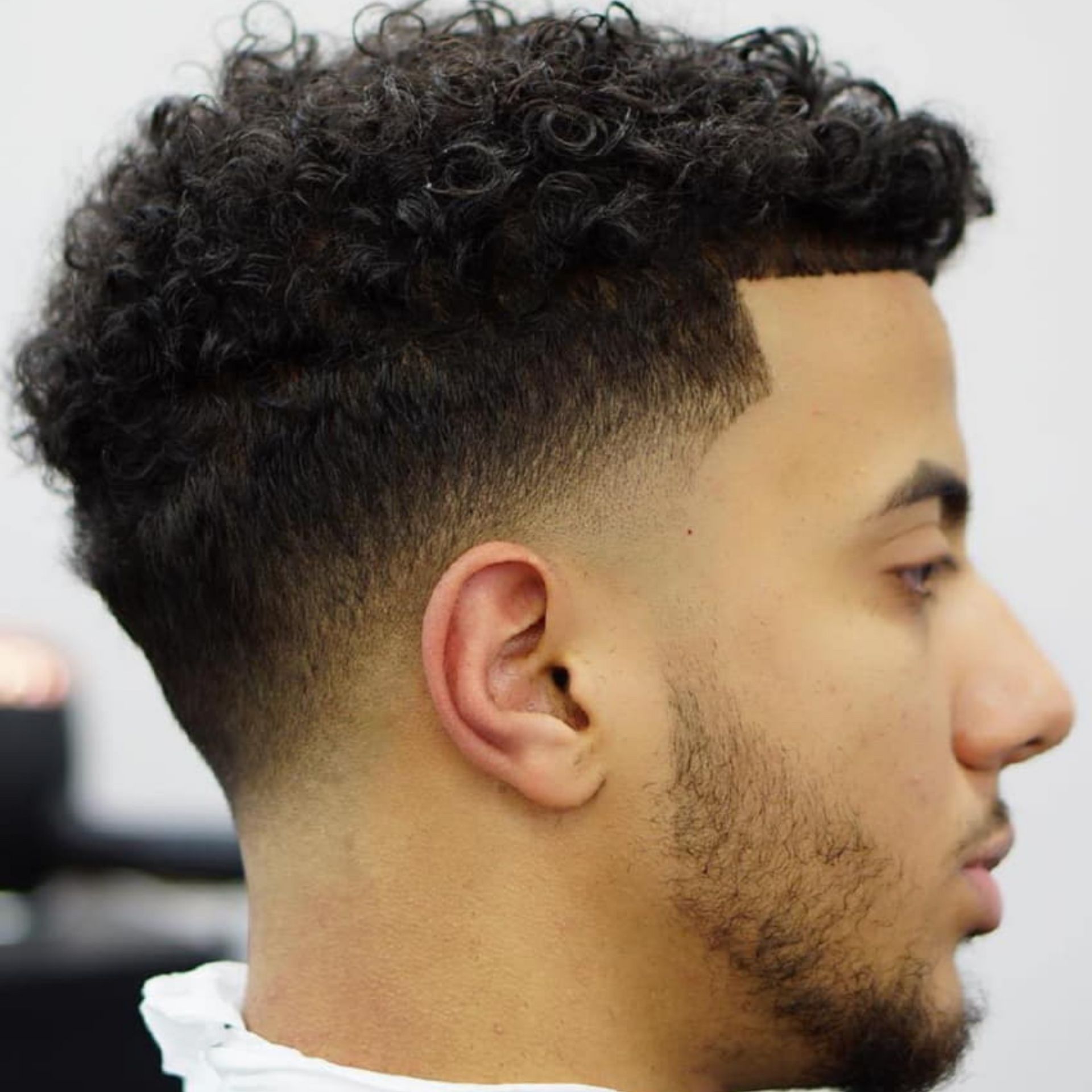 Perfil de un hombre con un corte de pelo rizado degradado y barba recortada, vestido con una camisa blanca.