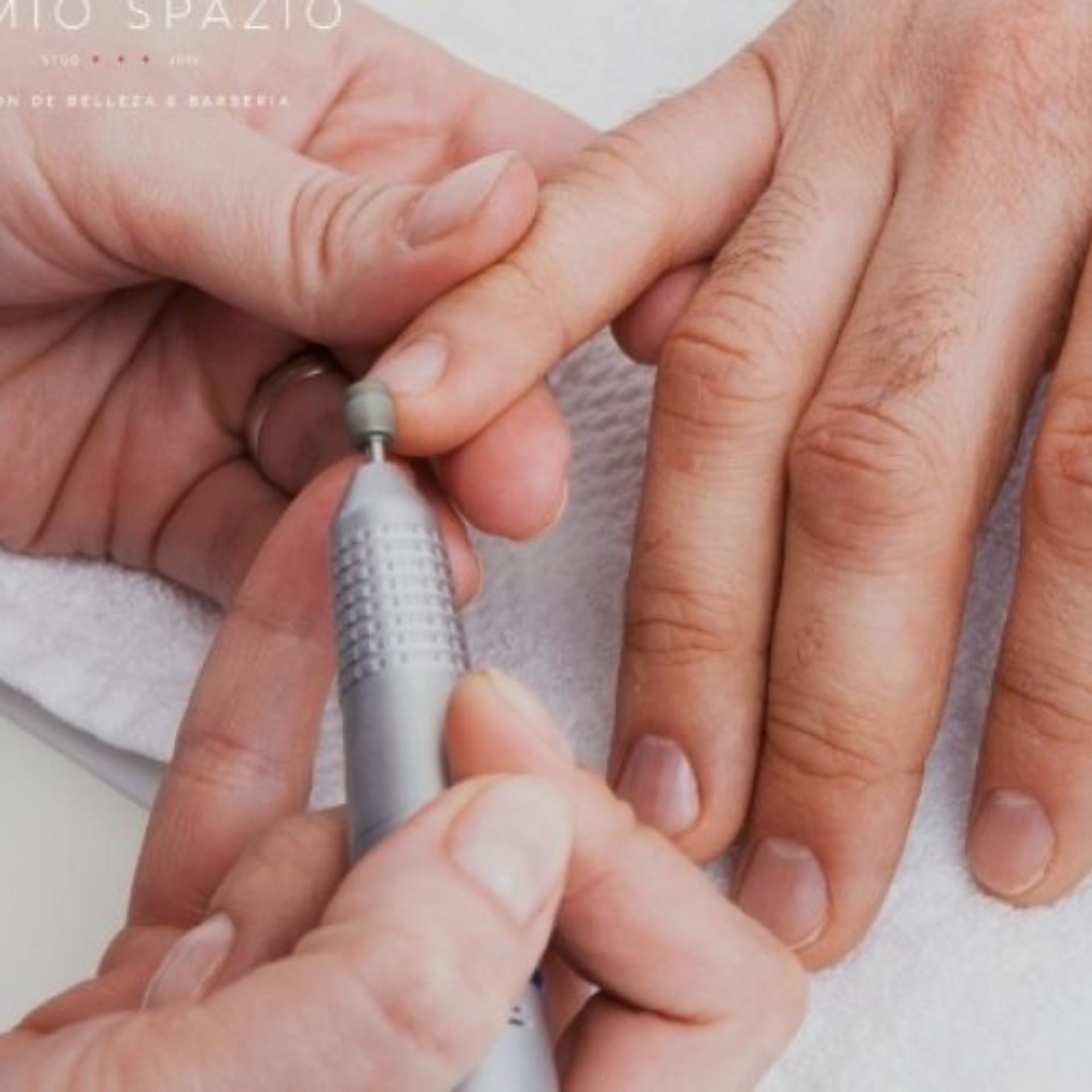Manos recibiendo una manicura con una herramienta de taladro de uñas sobre una toalla blanca.