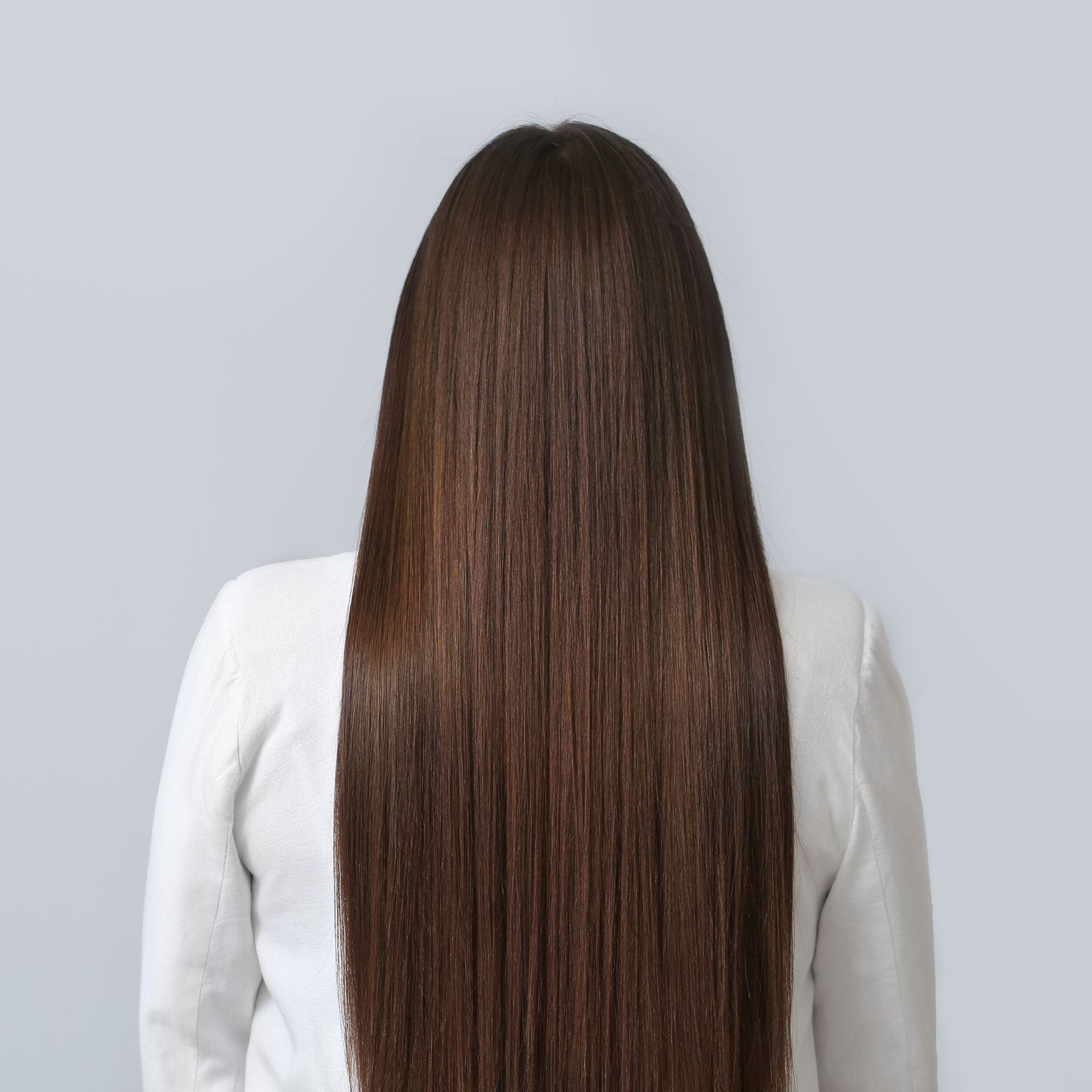 Vista posterior de una persona con cabello largo, liso y castaño, que lleva una blusa blanca sobre un fondo gris.