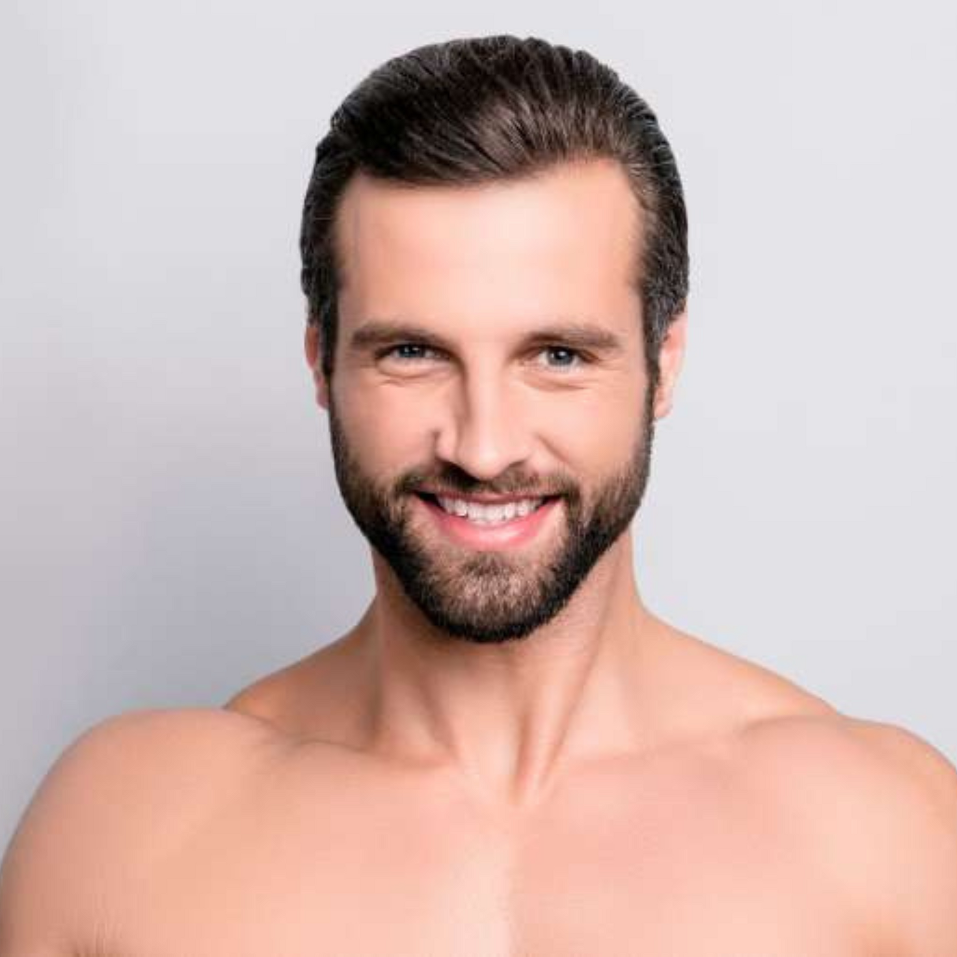 Hombre sonriente sin camisa, con cabello oscuro y barba, sobre un fondo gris claro.