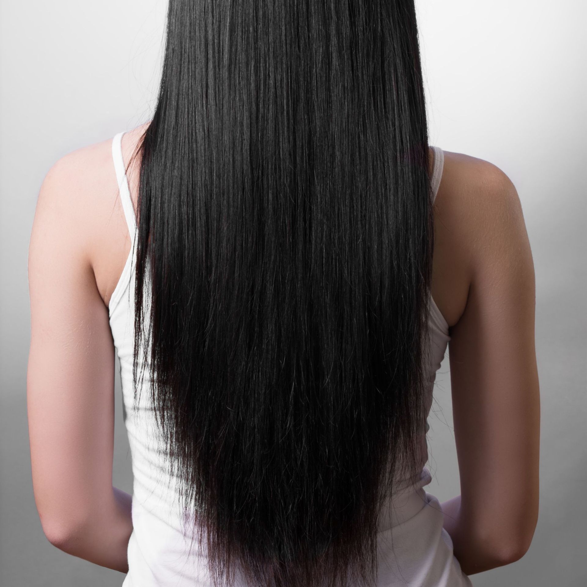 Persona con cabello largo, liso y negro, que lleva una camiseta blanca de tirantes, vista desde atrás.
