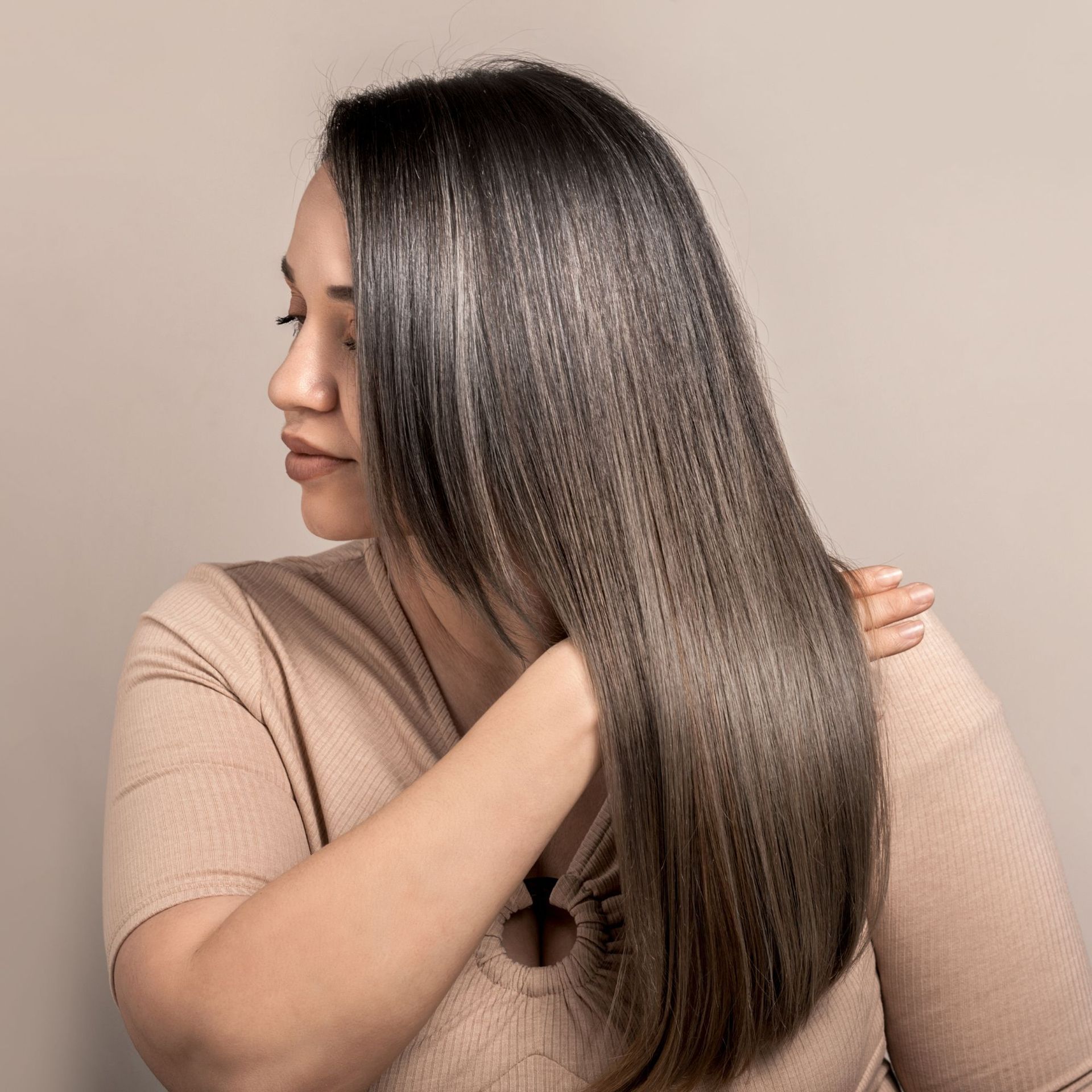 Mujer con cabello largo, liso y castaño, cepillándoselo contra un fondo beige.
