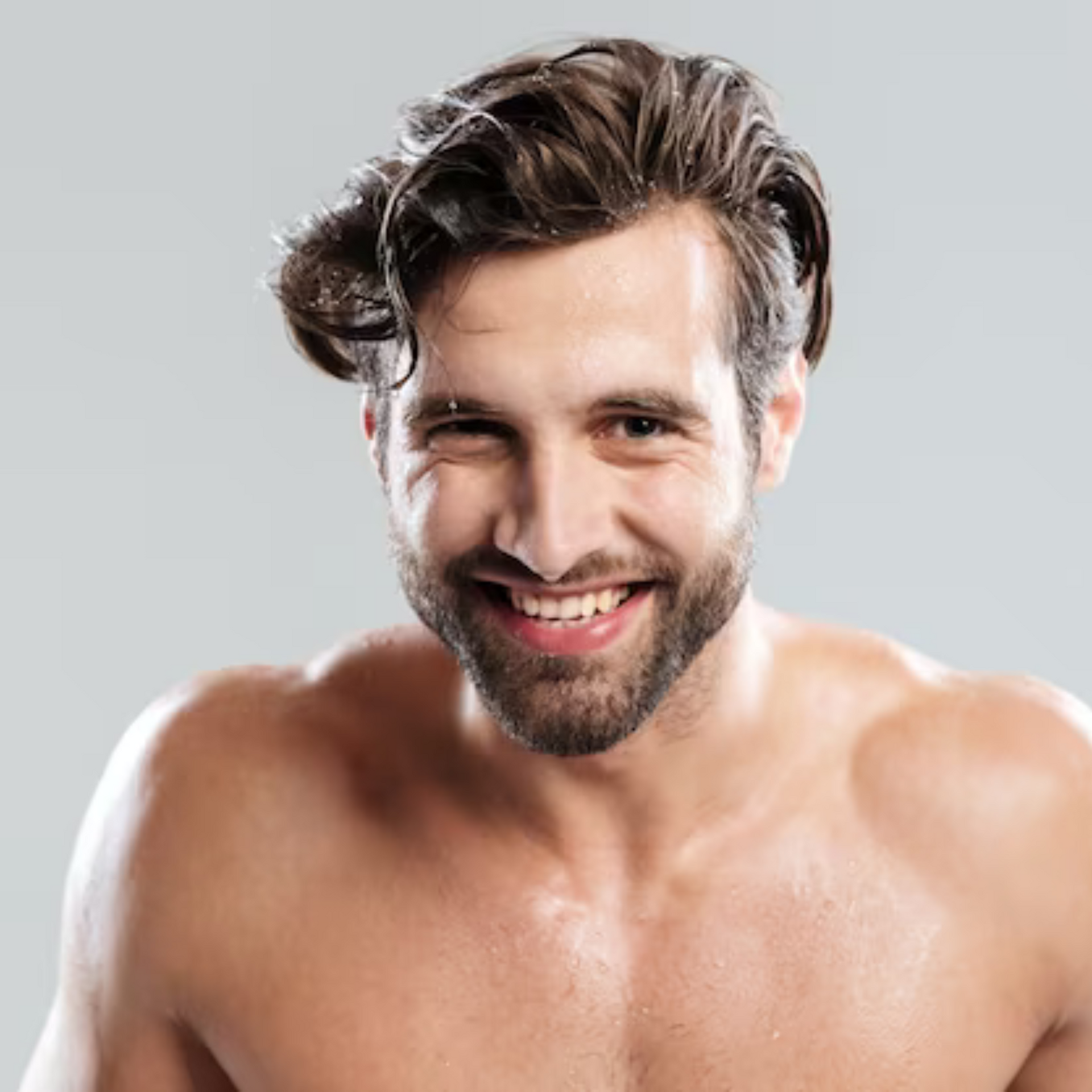 Hombre sonriente sin camisa, con cabello y barba despeinados, sobre un fondo gris claro.