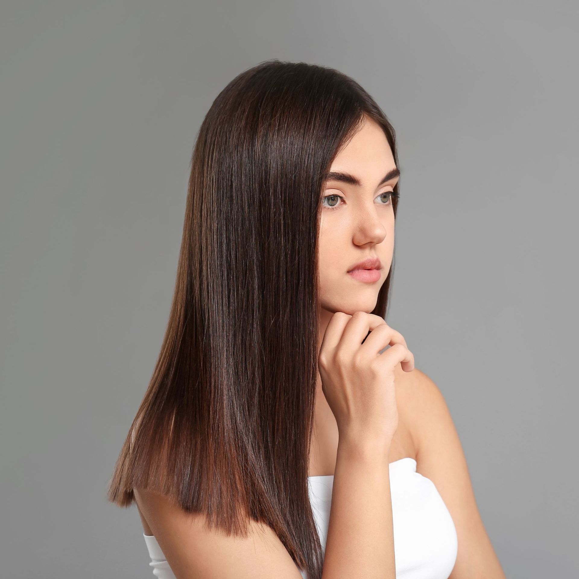 Mujer con cabello liso oscuro y blusa blanca, apoyando la barbilla en la mano sobre un fondo gris.