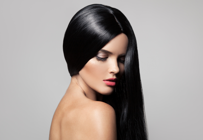 Mujer con cabello largo y negro y lápiz labial rosa, mirando hacia abajo contra un fondo gris.