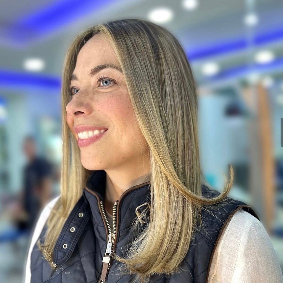 Persona sonriente con cabello rubio largo y chaleco azul marino, de pie en un interior con un fondo de oficina borroso.