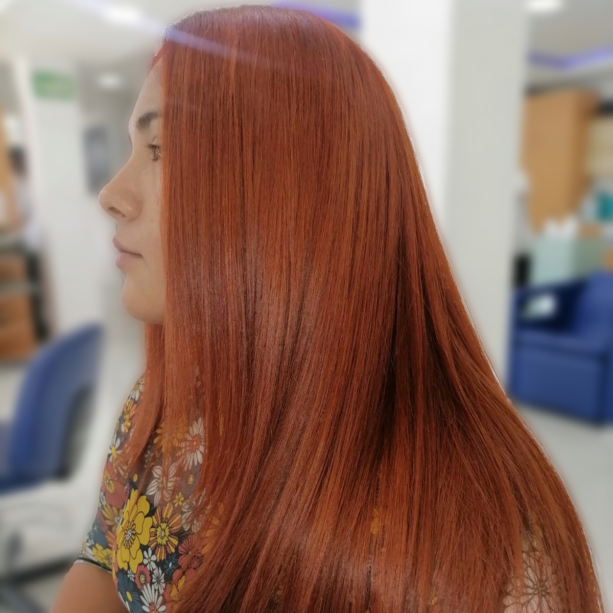 Perfil de una persona con cabello largo, liso y rojo en un entorno interior borroso.