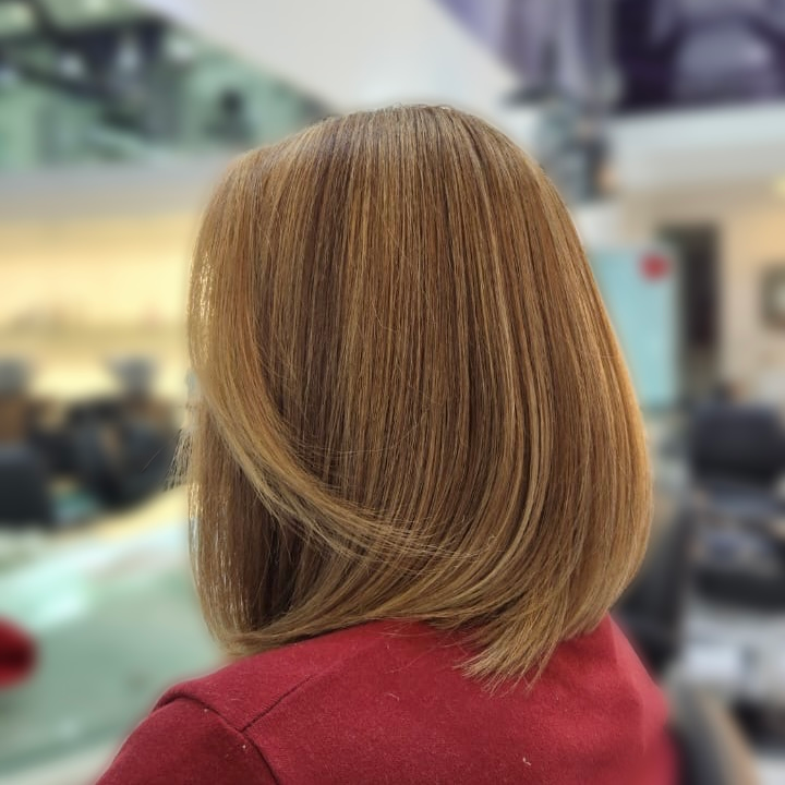 Vista trasera de una persona con cabello liso castaño claro, vestida con una chaqueta roja, en interiores con fondo desenfocado.