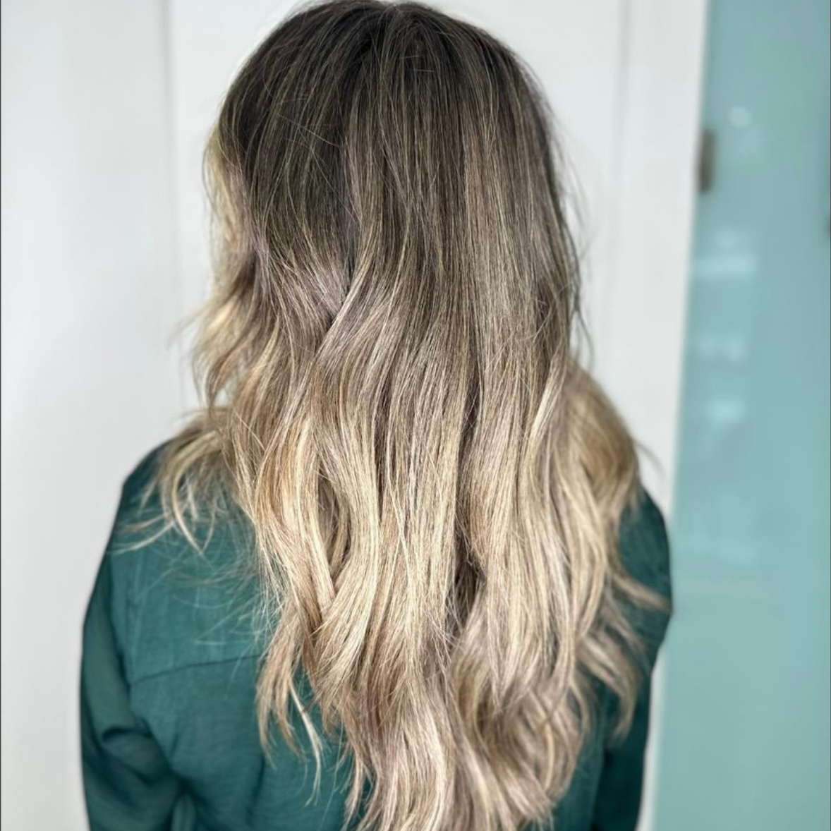 Vista posterior de una larga melena rubia ondulada con raíces oscuras sobre una blusa verde.