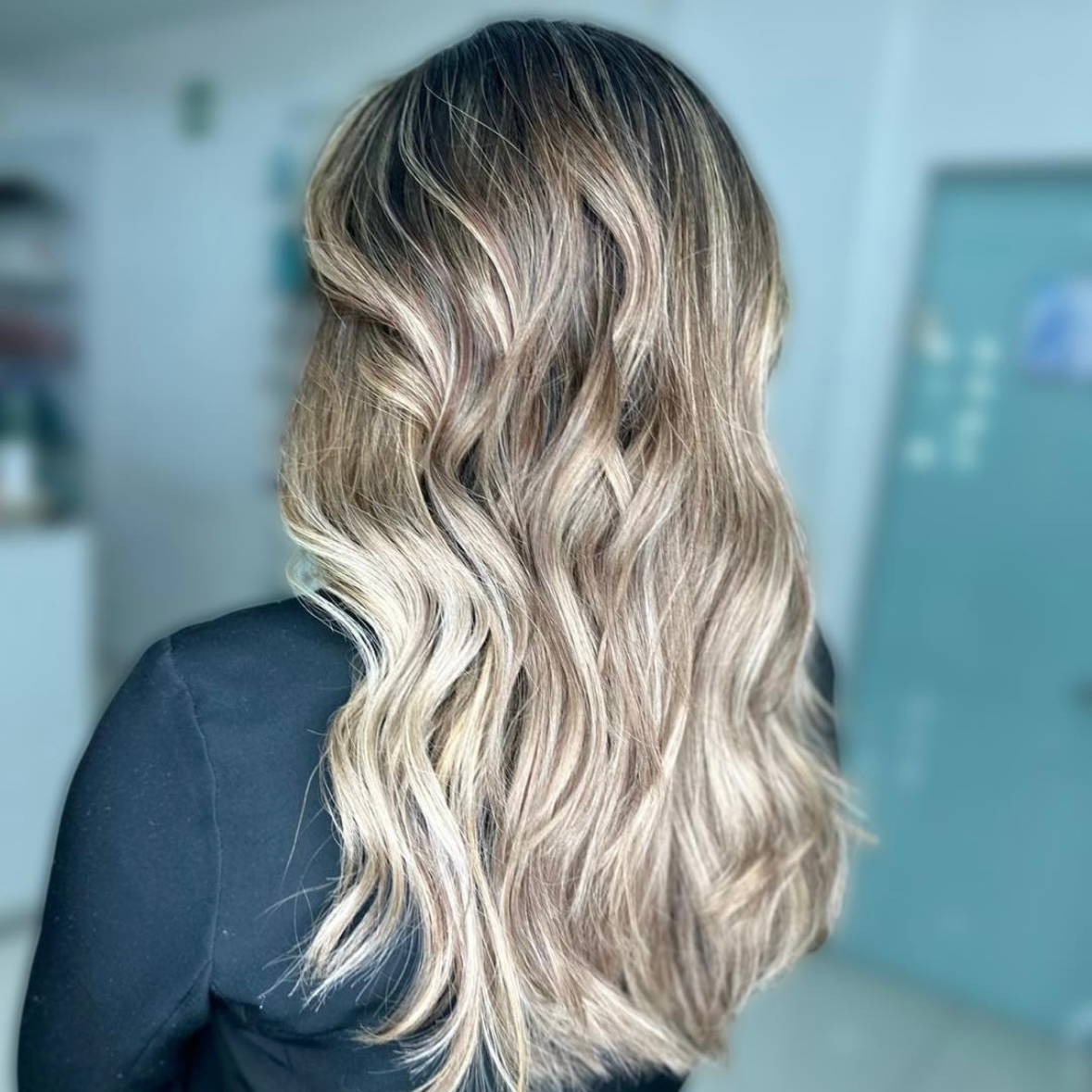 Cabello rubio ondulado con mechas balayage, peinado en ondas sueltas sobre un fondo interior desenfocado.