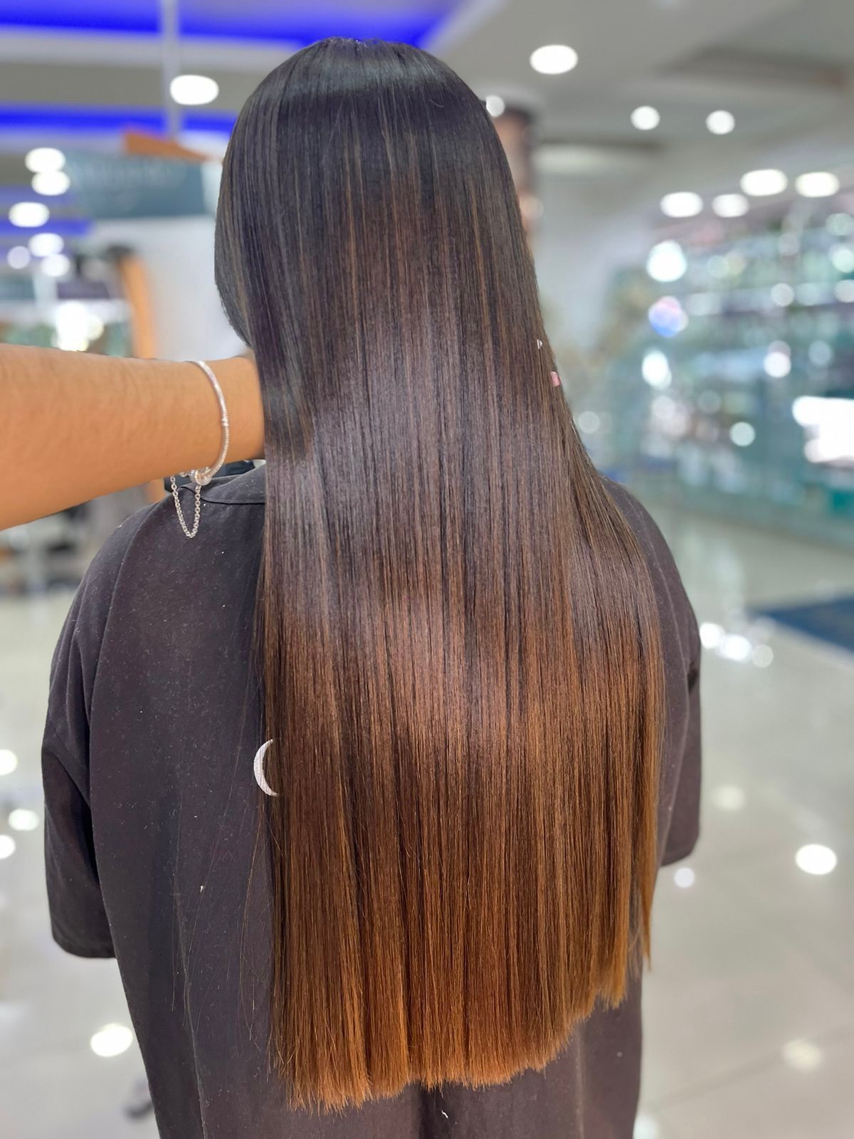 Cabello largo, liso y castaño con puntas más claras, visto desde atrás en un salón de belleza luminoso.