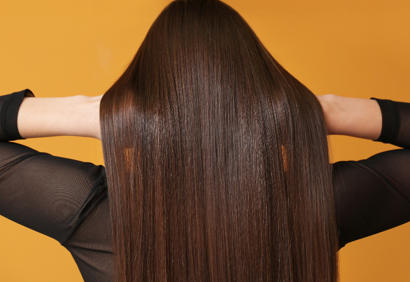 Persona con cabello castaño largo vista de espaldas, con las manos levantadas detrás de la cabeza sobre un fondo naranja.