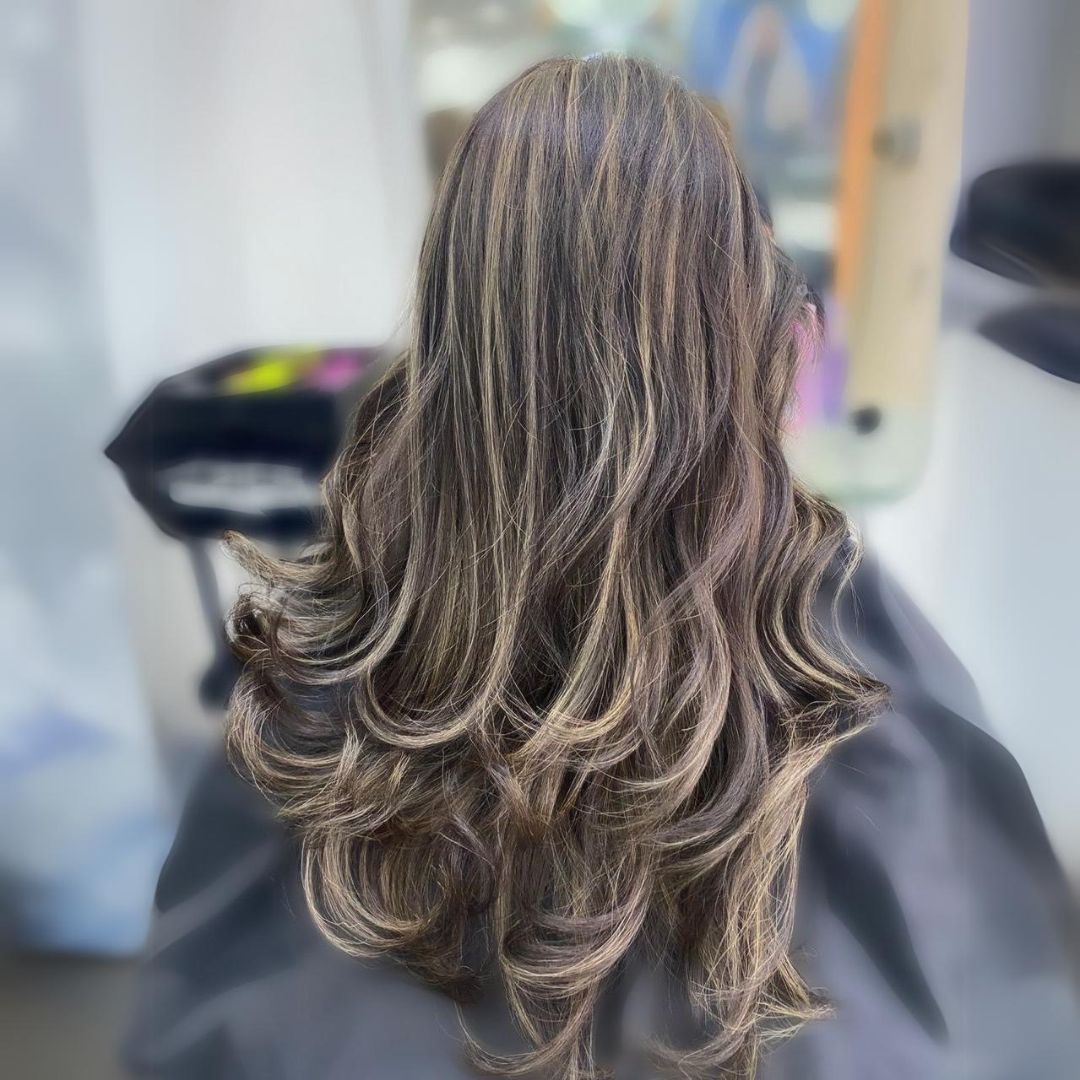Vista posterior de una larga melena castaña en capas con reflejos rubios en una silla de peluquería.