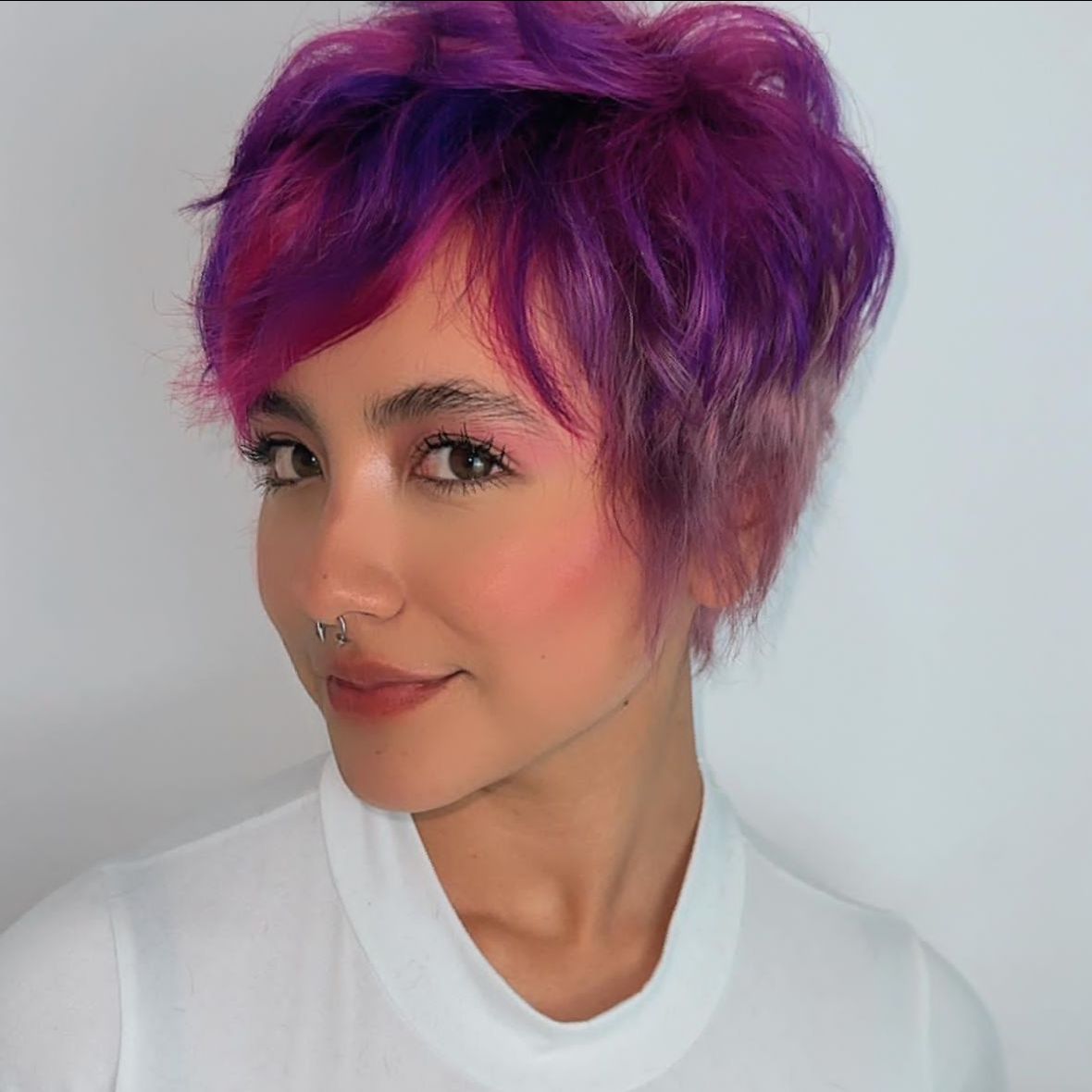 Retrato de una persona con cabello corto morado y camisa blanca, sonriendo sobre un fondo claro.