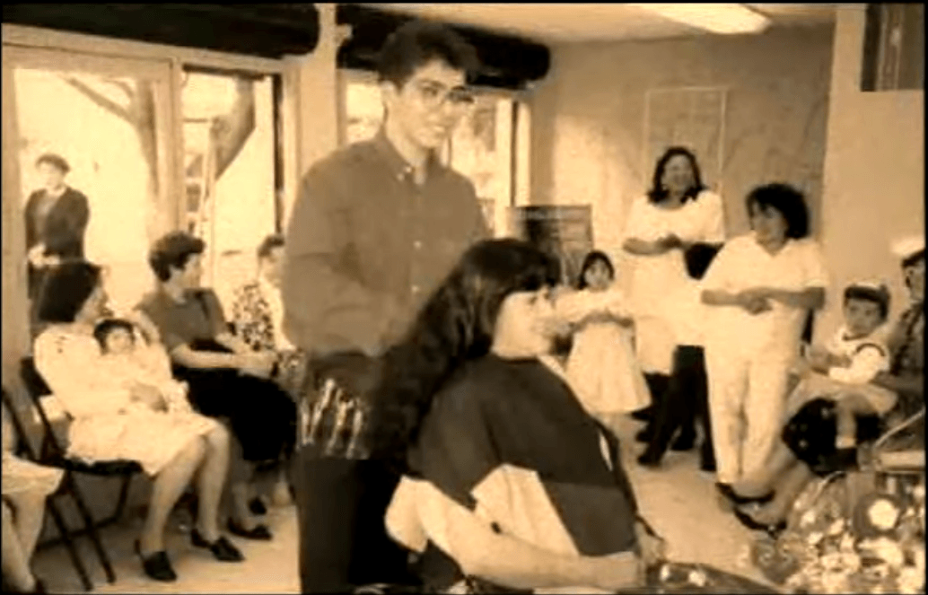 Escena de salón en tonos sepia con un hombre de pie junto a una mujer sentada, rodeados de espectadores.