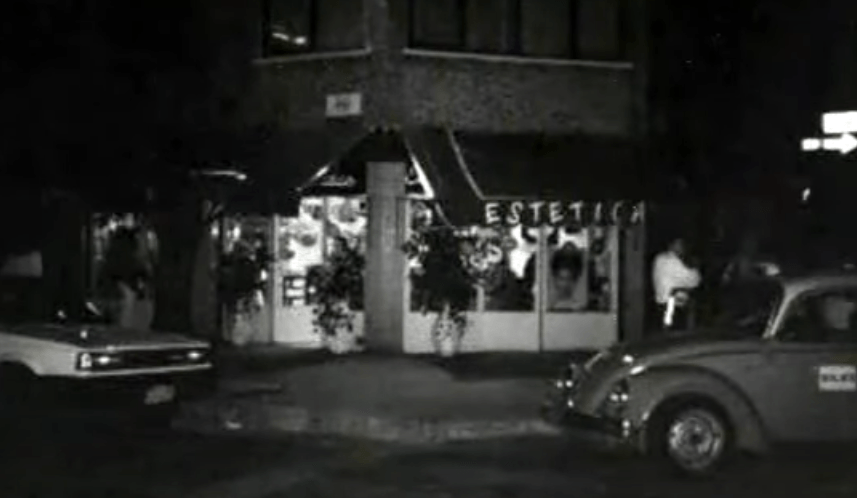 Escena callejera en blanco y negro con coches aparcados frente a una tienda por la noche; la gente se había congregado cerca de la entrada.