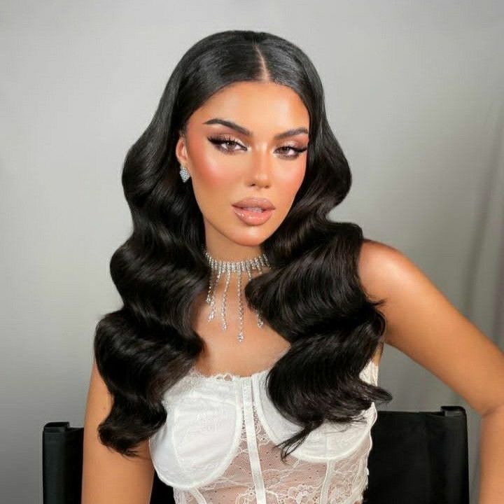 Mujer con cabello largo, ondulado y oscuro, vestida con un corsé blanco y una gargantilla enjoyada, posando sobre un fondo gris.