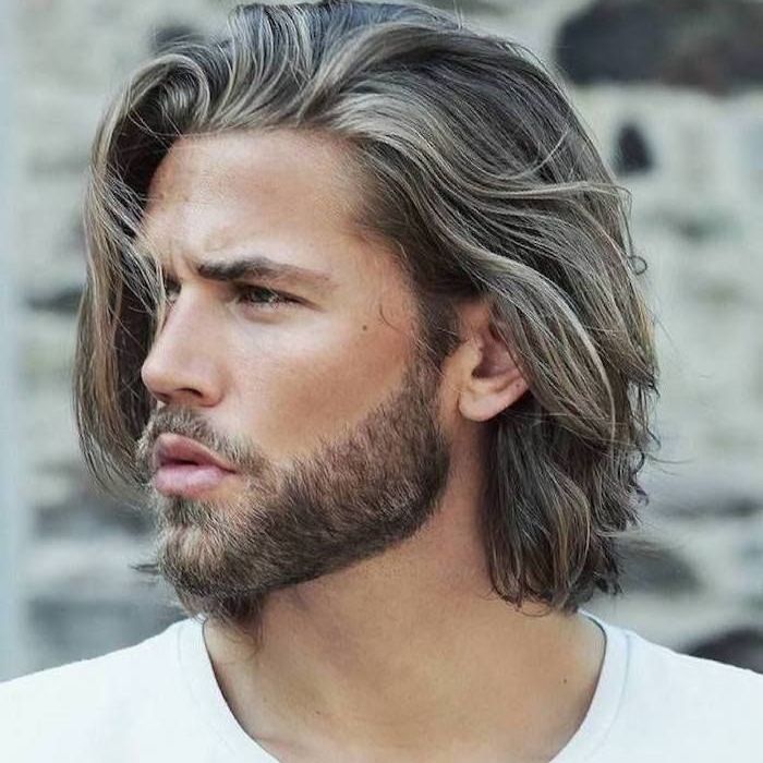Retrato en primer plano de un hombre con cabello gris ondulado y barba, mirando hacia la izquierda contra un muro de piedra borroso.