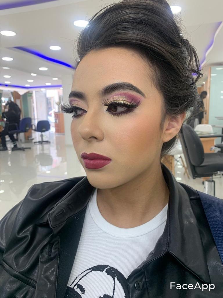 Persona con maquillaje llamativo y peinado elaborado en un salón luminoso, vestida con una chaqueta negra y una camisa blanca.