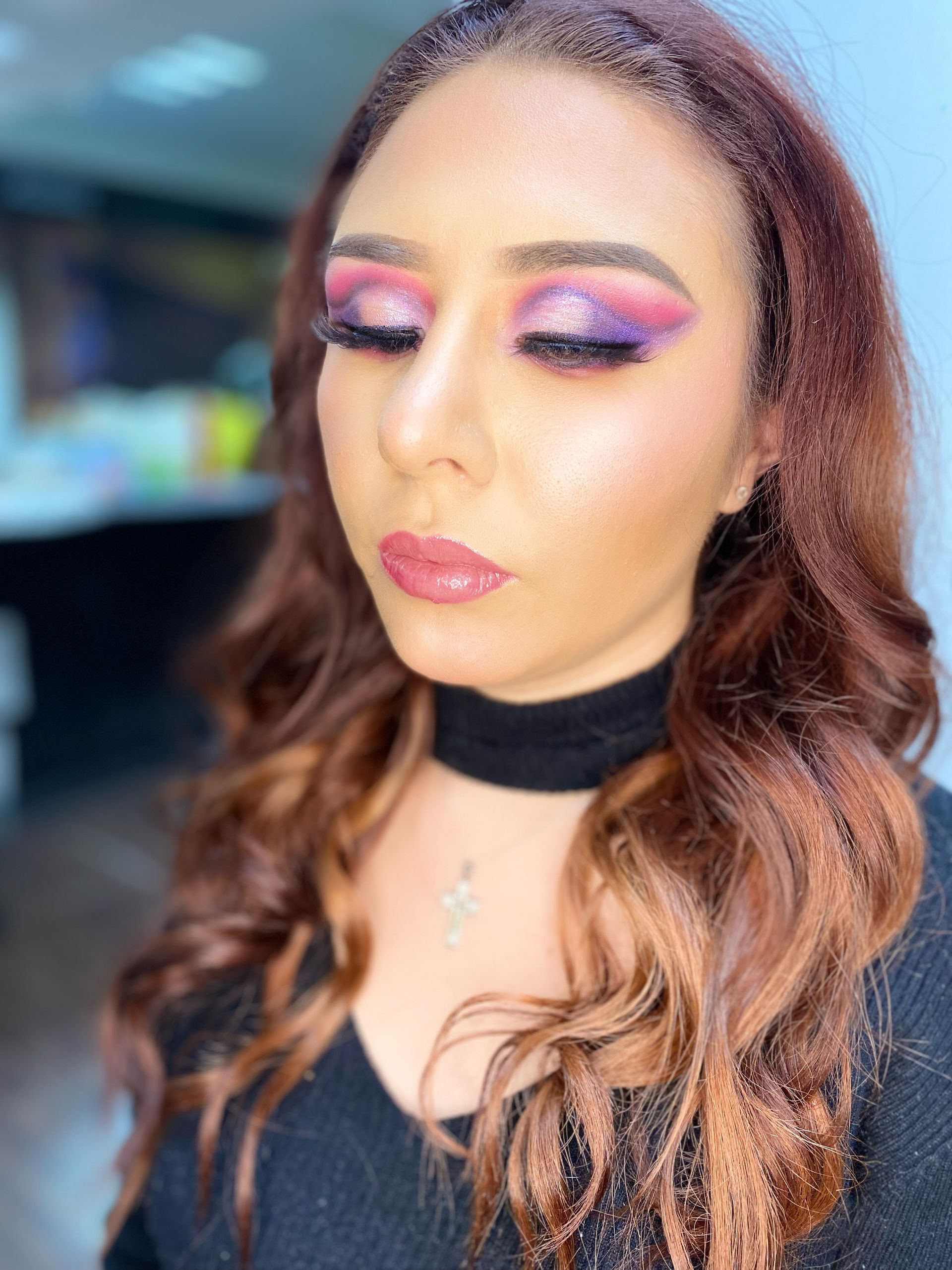 Retrato de una mujer con maquillaje de ojos rosa y morado, labios brillantes y cabello castaño ondulado sobre un fondo desenfocado.