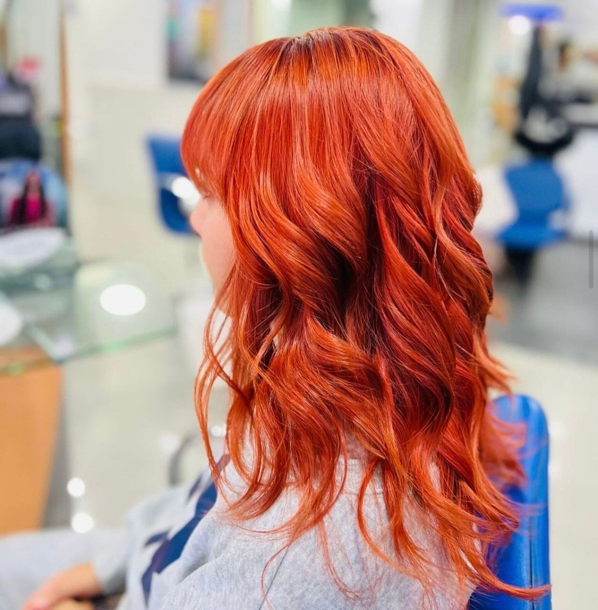 Vista lateral de una persona con cabello ondulado de color rojo brillante sentada en una silla de peluquería, vistiendo una chaqueta vaquera.