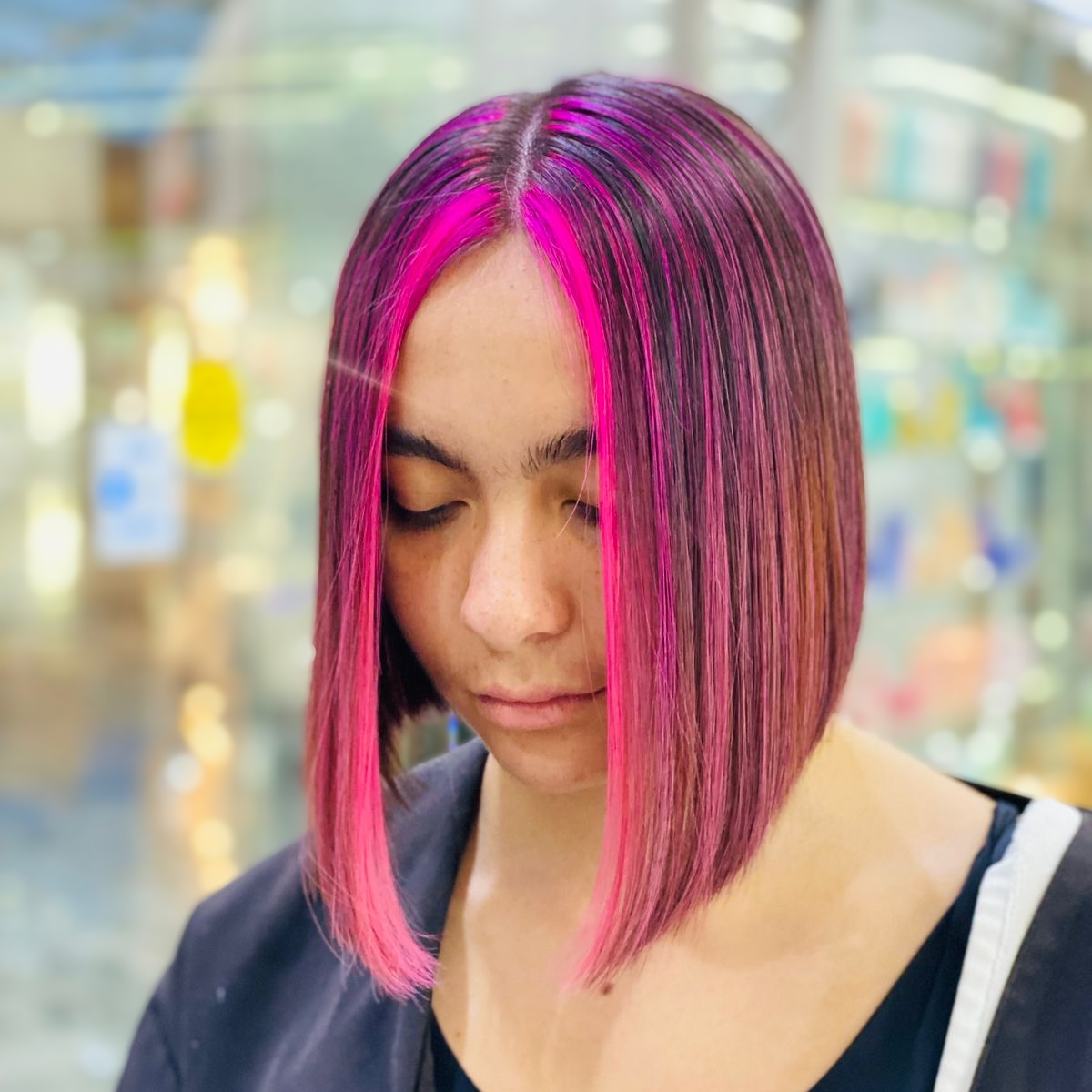 Persona con el pelo liso de corte bob teñido de rosa brillante, mirando hacia abajo en un entorno interior suavemente desenfocado.