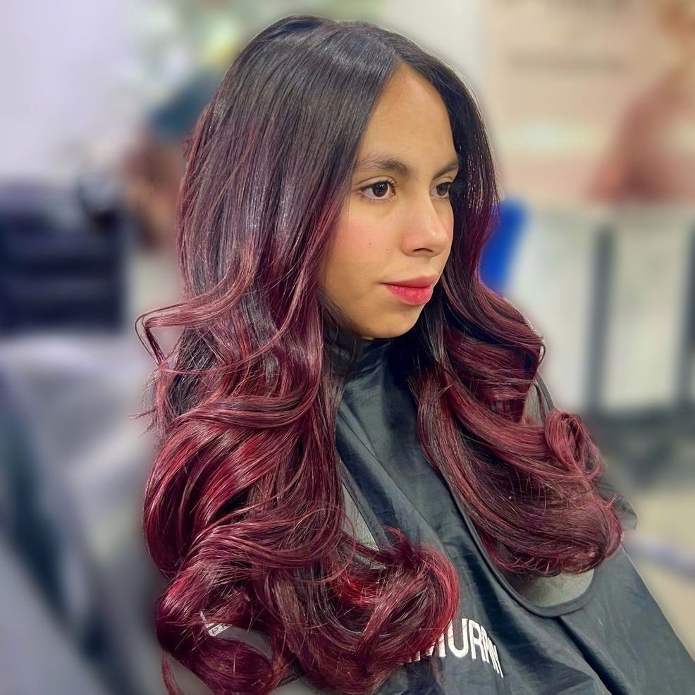 Mujer con cabello largo y ondulado color burdeos sentada en una silla de peluquería, con una capa negra, mirando hacia un lado.