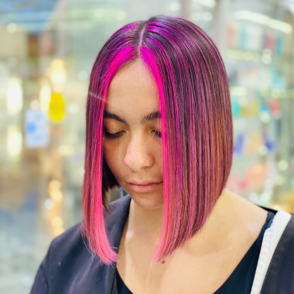 Persona con corte de pelo bob liso teñido de rosa y morado, mirando hacia abajo en un escenario de salón borroso.