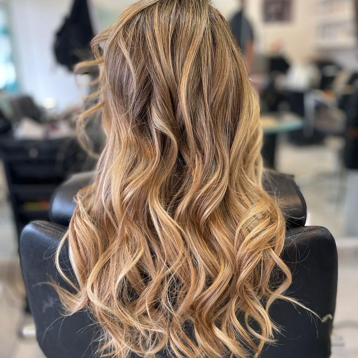 Cabello rubio largo y ondulado con reflejos, peinado en rizos sueltos, visto desde atrás en una silla de peluquería.