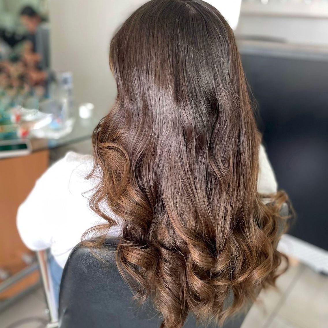 Mujer con cabello castaño largo y ondulado vista de espaldas en una silla de peluquería.