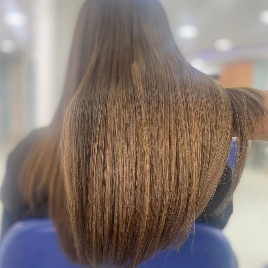 Vista trasera de una persona con cabello largo, liso y castaño, sentada en una silla azul, en un ambiente interior con iluminación tenue.