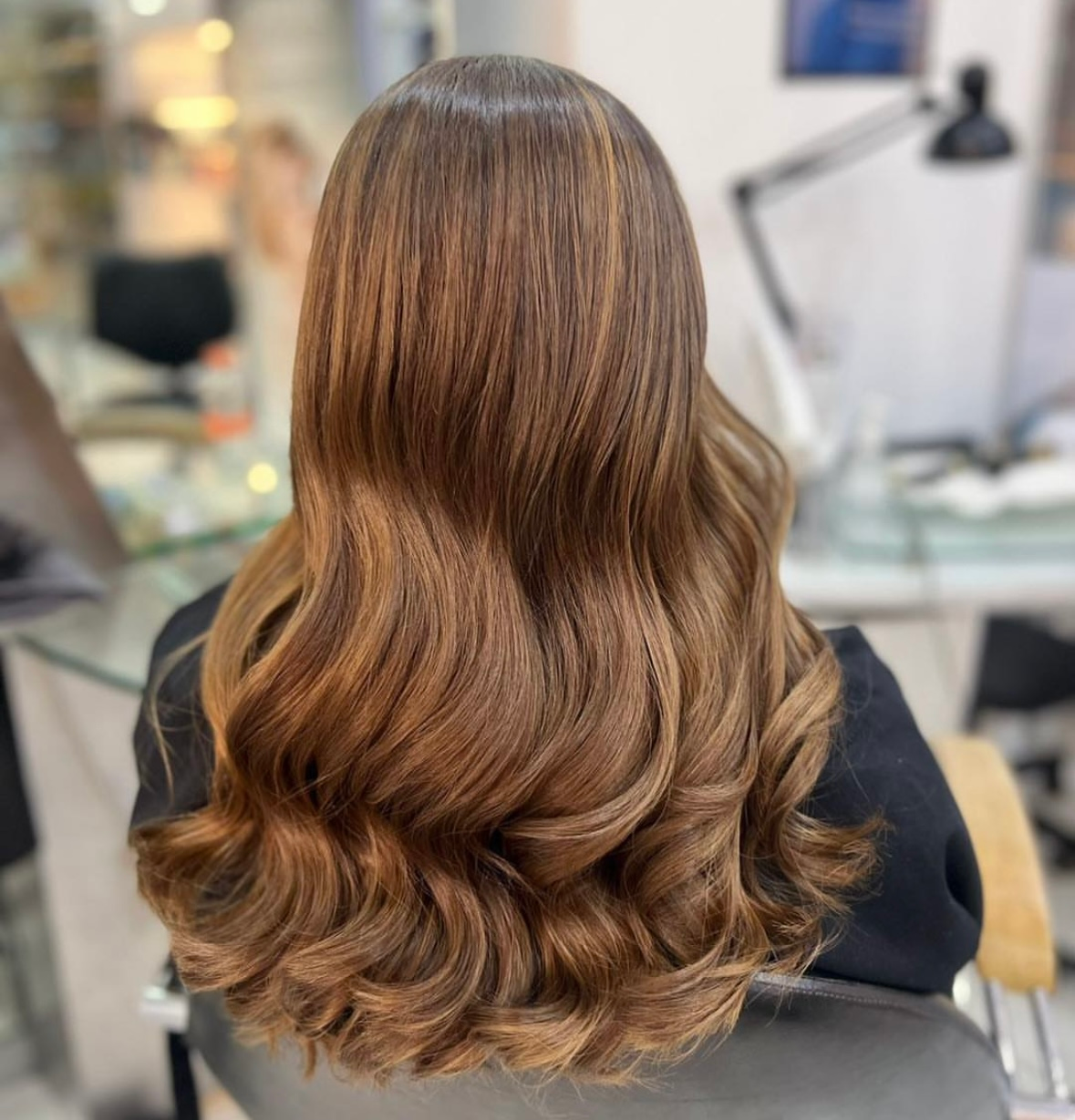 Vista posterior de una larga melena castaña ondulada en una silla de peluquería, con rizos brillantes y capas.