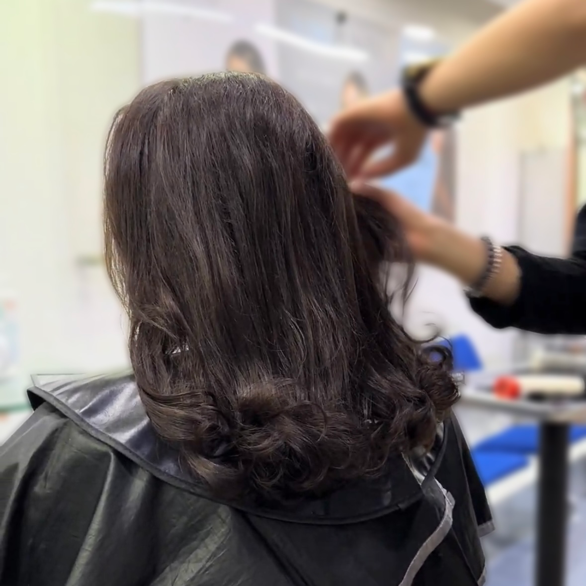 Una persona con cabello oscuro se está cortando el pelo en una peluquería, mientras un estilista le está peinando.