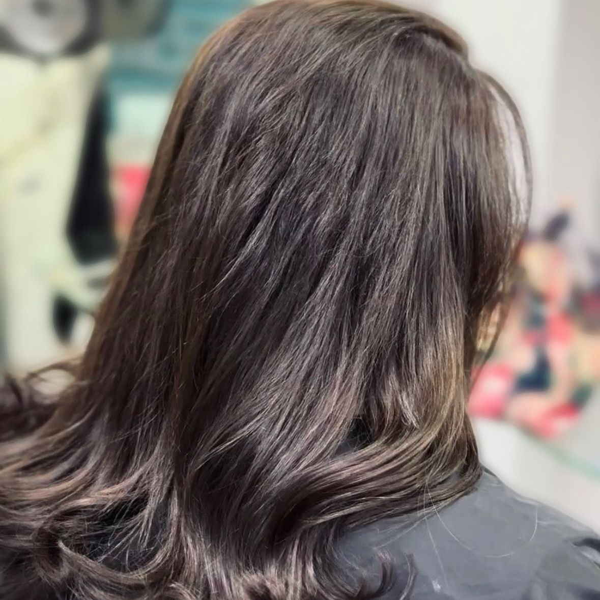 Espalda de una persona con cabello oscuro hasta los hombros sentada en una silla de peluquería.