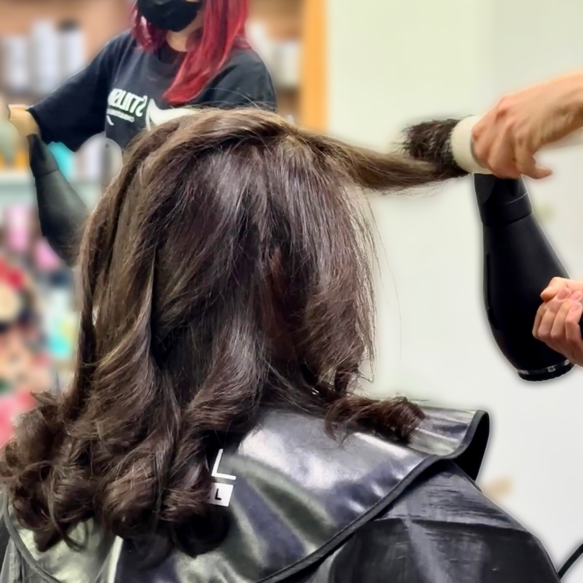 Una persona se está secando el pelo con secador en una silla de peluquería, vistiendo una capa negra.