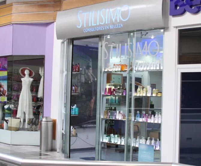 Fachada de un salón de belleza con ventanas de cristal, expositores de productos y un maniquí con un vestido blanco.