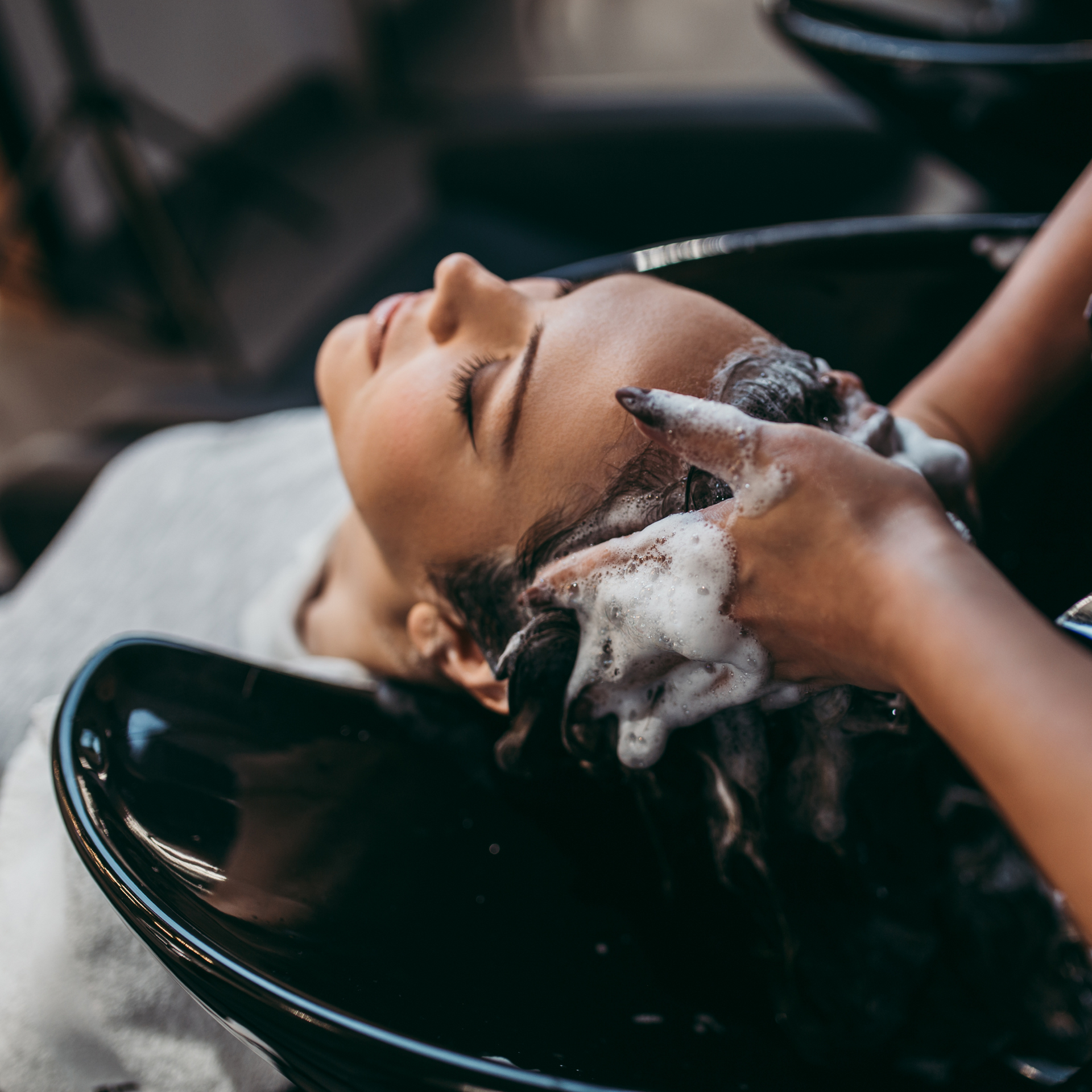 Peluquera lavando el cabello de una clienta en un lavabo negro de salón con espuma de champú blanca.